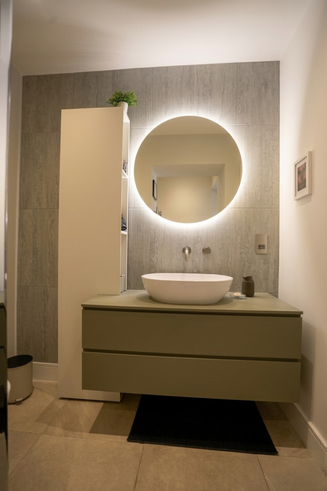 Contemporary bathroom design with backlit round mirror, countertop basin on a wall-hung vanity unit and fluted stone-effect feature wall, Islington London