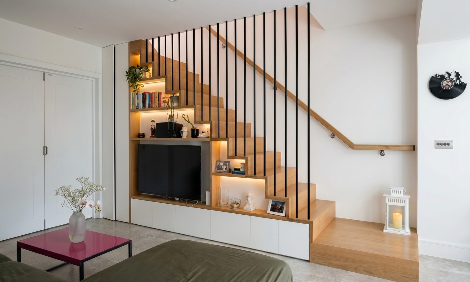 Bespoke oak staircase with black vertical balustrade and integrated joinery storage wall featuring TV, shelving and recessed LED lighting, Islington London