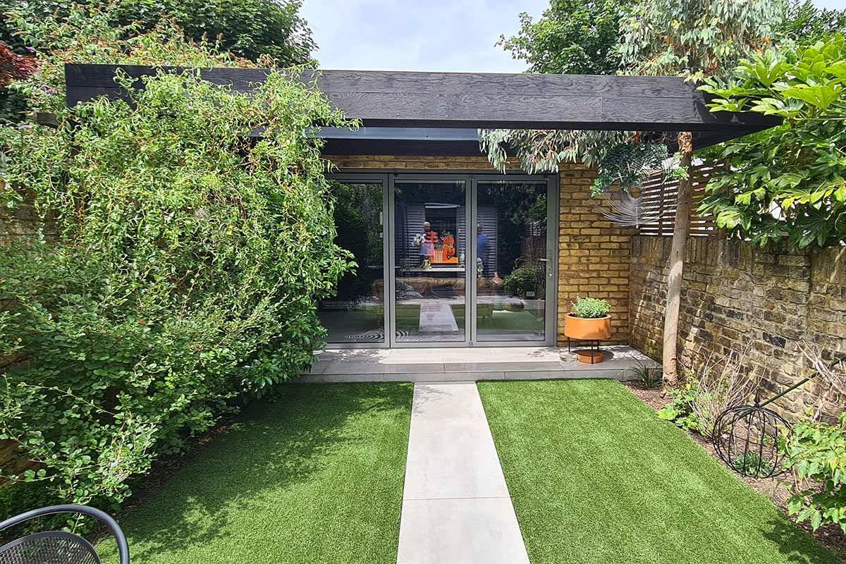 View from the garden of a contemporary garden room with open aluminium bifold doors, black timber roof fascia and London stock brick walls, glimpsing the interior with leather sofa, London