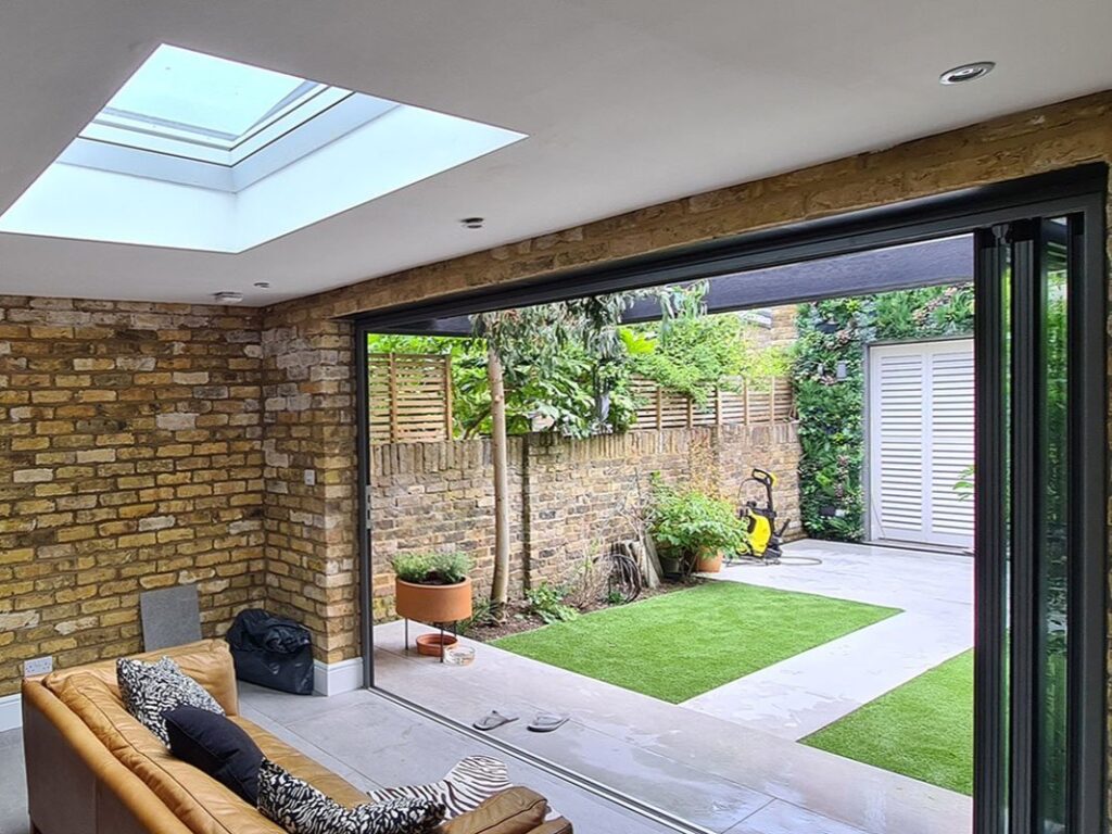 Garden room lounge interior with black feature wall, tan leather sofa, exposed London stock brick side walls and skylight overhead, London