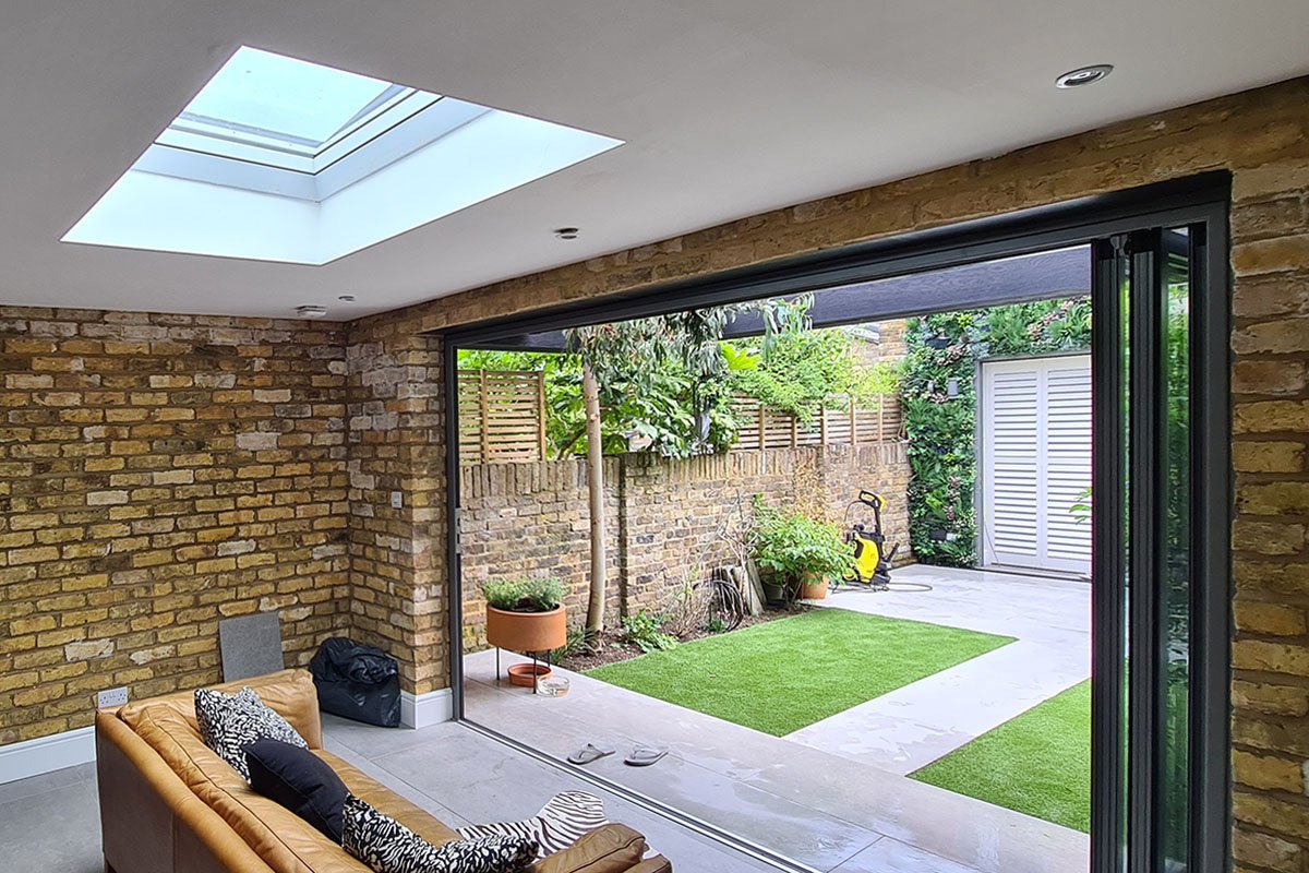 Garden room lounge interior with black feature wall, tan leather sofa, exposed London stock brick side walls and skylight overhead, London