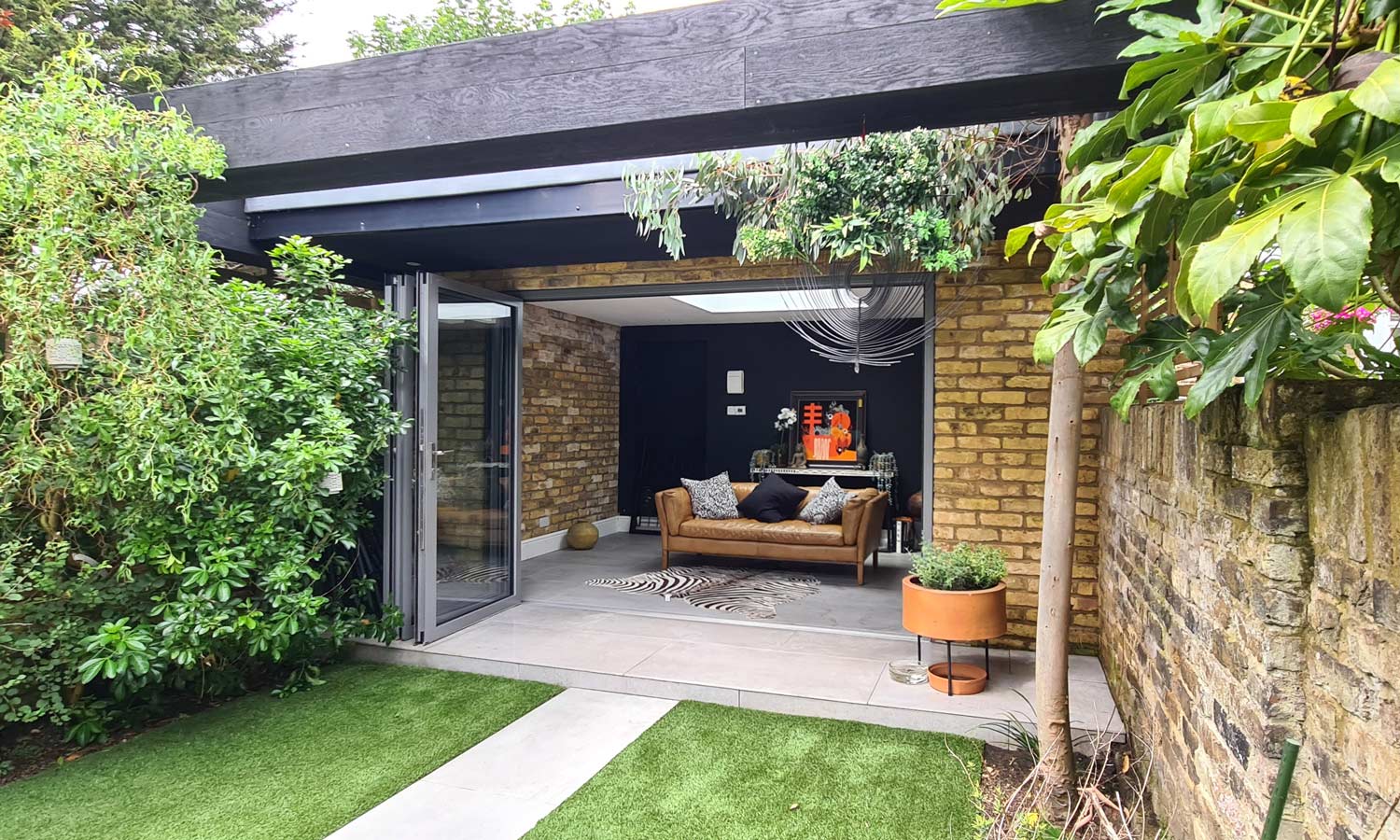 Architectural detail looking up at a garden room's cantilevered black timber roof, with overhanging foliage and steel beam against the sky, London