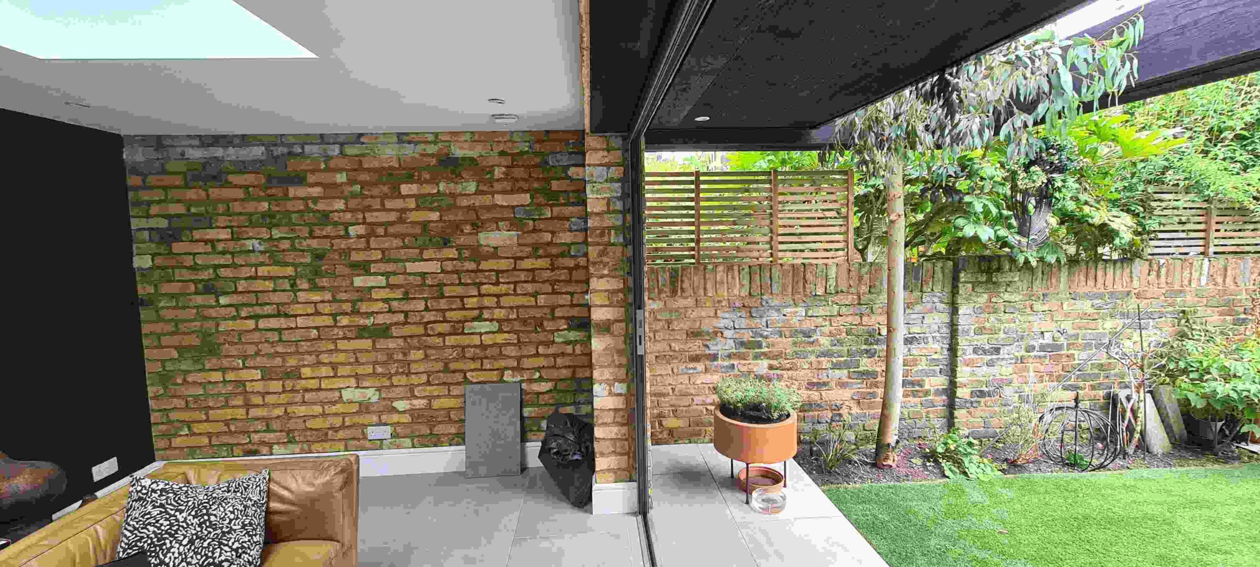 Garden room interior with skylight and open bifold doors revealing a small patio with bistro furniture and lawn, exposed London stock brick wall, London