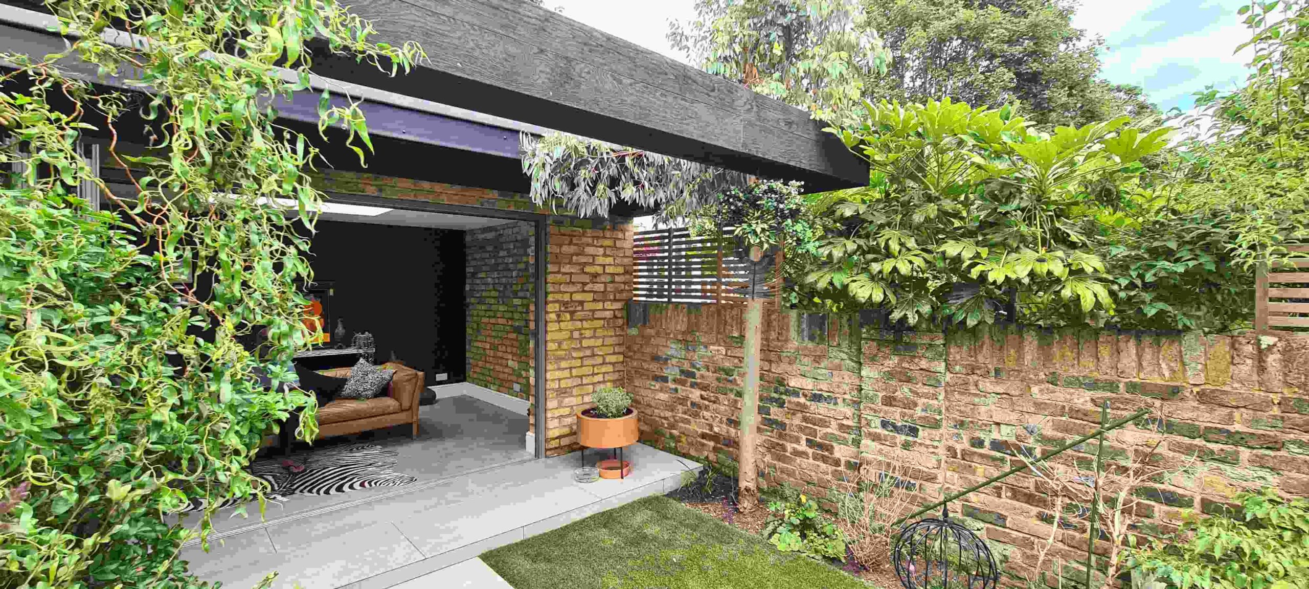 Panoramic view of a garden room blending interior and exterior, with exposed London stock brick walls, black accents and bifold doors opening to a planted urban garden, London