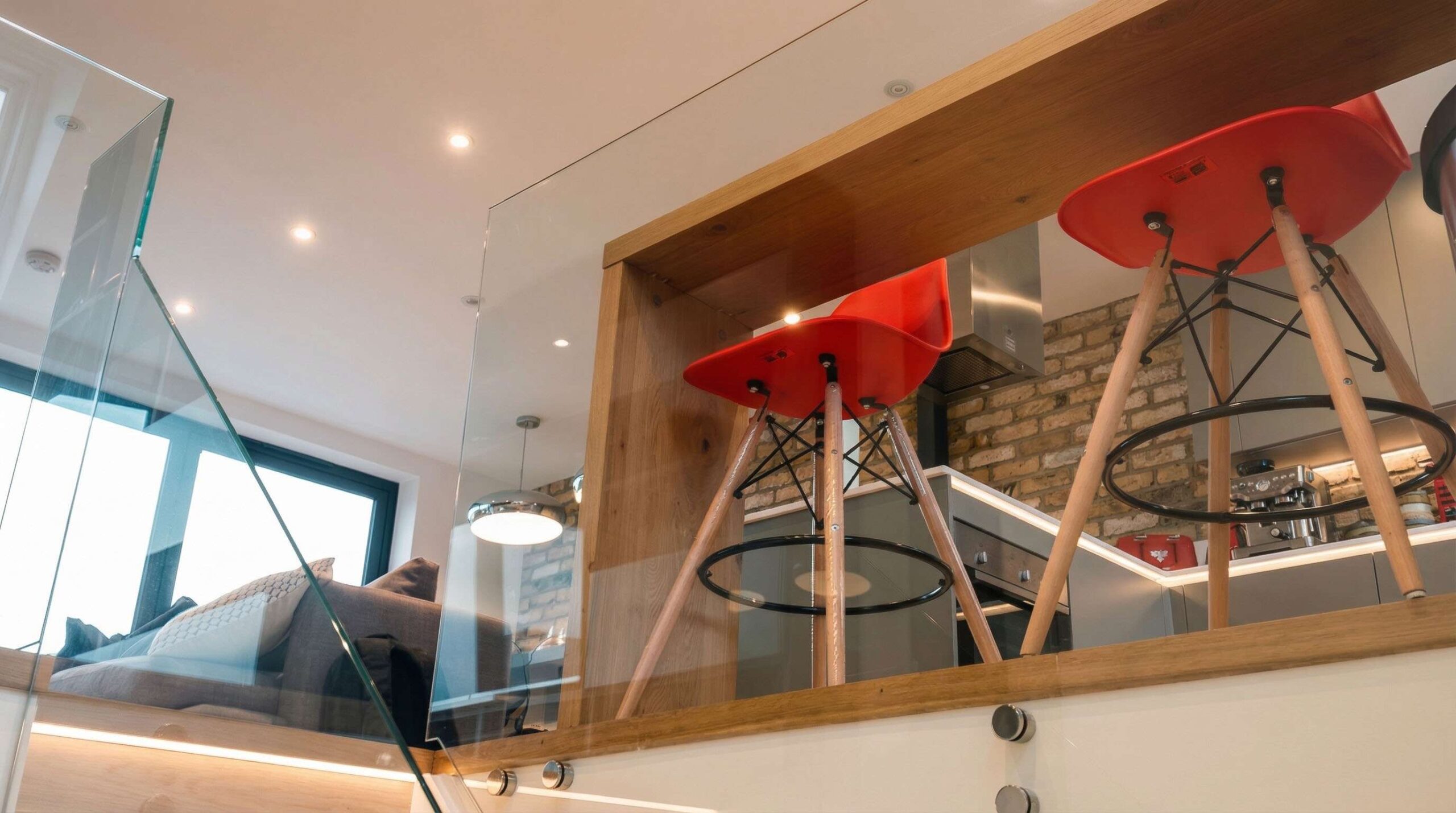 Split-level apartment interior with frameless glass balustrade, oak handrail and Eames-style bar stools overlooking the kitchen below, Fulham London