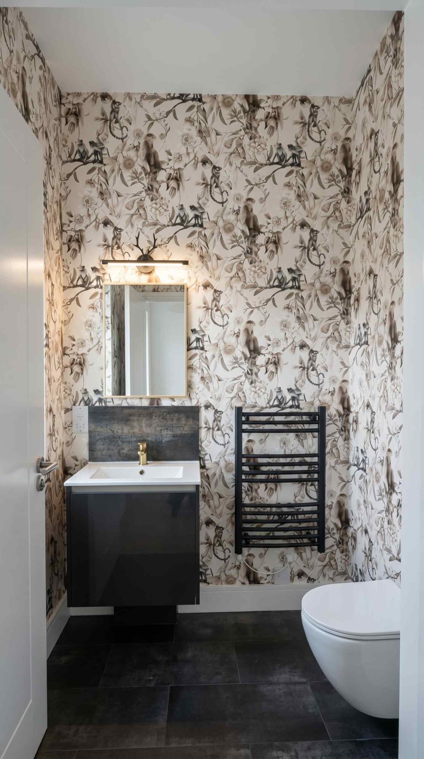 Statement bathroom with tropical wildlife wallpaper, gloss black vanity unit, brass tap, wall-hung WC and black towel radiator in a period property, Islington London